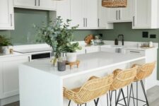 a welcoming farmhouse kitchen with white cabinets and a green backsplash, stone countertops, a white kitchen island, rattan stools and woven lamps
