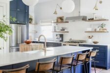 an airy kitchen with bold blue lower cabinets, a large kitchen island, white countertops, open shelves, pendant lamps and brass sconces
