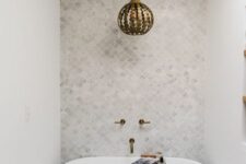 an elegant neutral bathroom with marble tiles, a small tub, a vanity and a wooden shelf, a whimsical pendant lamp