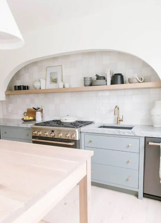 a neutral and airy kitchen with grey cabinets, a large arched niche with an open shelf used for displaying porcelain and a table that is a kitchen island
