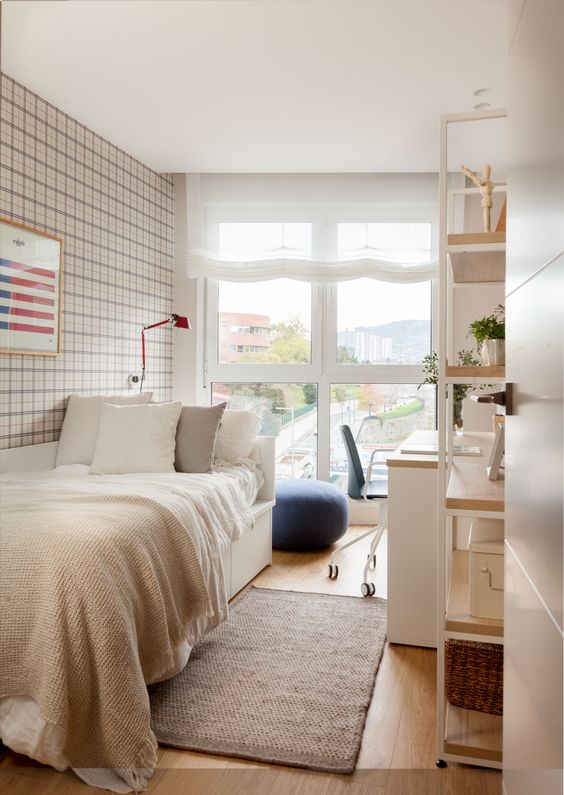 a small and functional guest bedroom with a floor to ceiling window, a bed with pillows, a desk, a chair and an open shelving unit