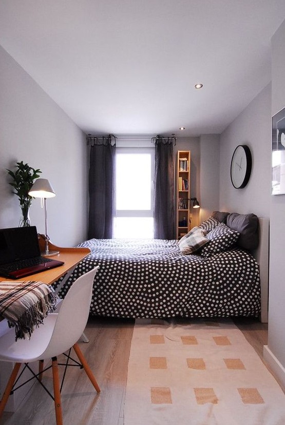 a compact bedroom with an upholstered bed and printed bedding, a wall-mounted desk and a white chair and grey curtains