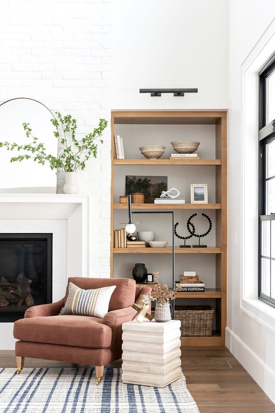 a modern farmhouse living room with a fireplace, built-in niche shelves of wood with decor, a burgundy chair and a side table