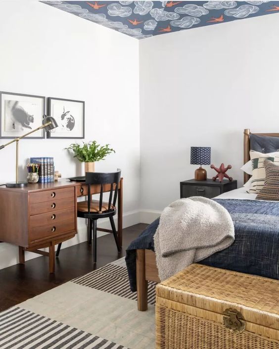 a guest bedroom with a bed and nightstands, a wicker chest for storage, a small stained desk and a black chair and some art