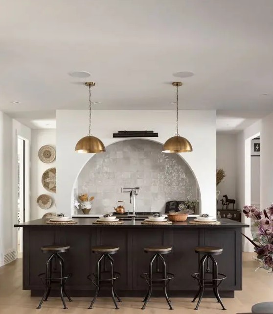 a modern kitchen with an arched niche clad with Zellige tiles and built-in cabinets, a black kitchen island, brass pendant lamps and industrial stools