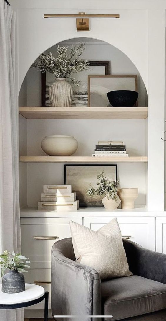 a neutral living room with an arched niche with books and lovely decor, a grey chair with books and greenery is amazing