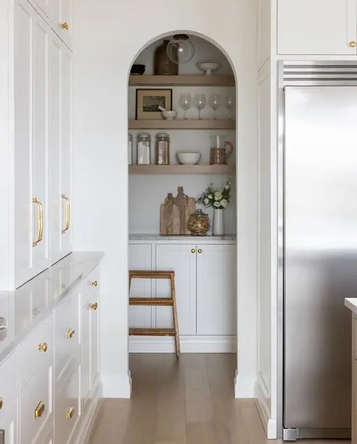 a white modern farmhouse kitchen with an arched niche that hides a pantry with cabinets and open shelves, a smart solution to save some pace