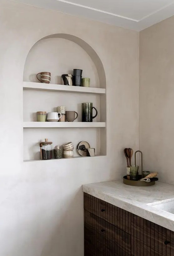 a contemporary kitchen with plaster walls, dark-stained cabines and neutral stone countertops, an arched niche for displaying mugs