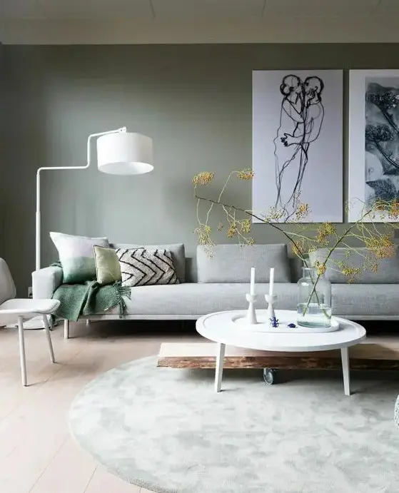a Scandinavian living room with a green accent wall, a grey long sofa with pillows, some artwork and some white furniture and a lamp
