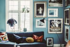 a Scandinavian living room with blue walls, a free form gallery wlal, a black sofa, an amber leather chair and a glass coffee table
