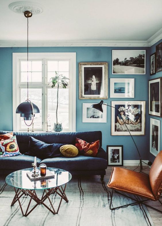 a Scandinavian living room with blue walls, a free form gallery wlal, a black sofa, an amber leather chair and a glass coffee table