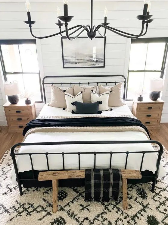 a catchy modern farmhouse bedroom with a wrought bed with contrasting bedding, a printed rug, stained nightstands, a chandelier