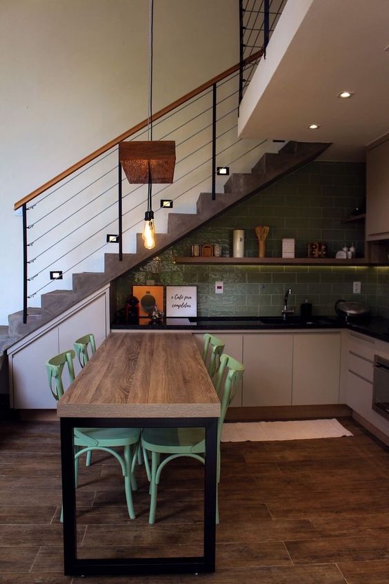 a catchy modern kitchen placed under the staircase, with a green tile backsplash, greige lower cabinets, black countertops and an open shelf