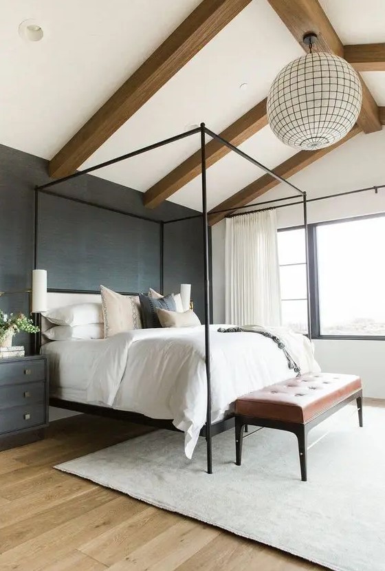 a chic modern farmhouse bedroom with wooden beams, a black grasscloth wallpaper wall, a black frame bed, black nightstands and a leather bench
