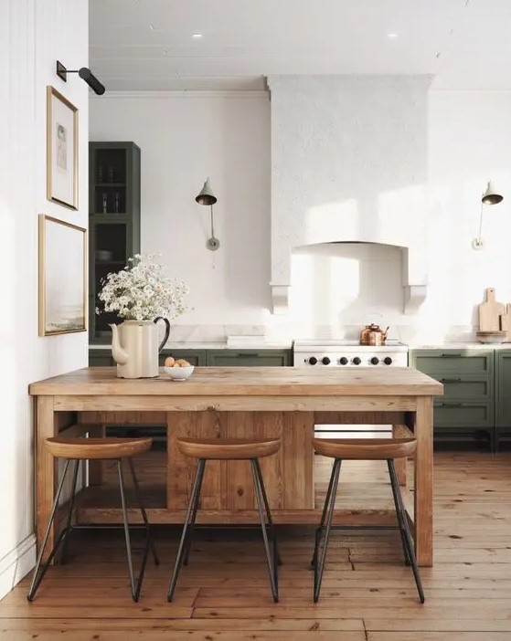 a chic modern farmhouse kitchen with green cabinets, a large white hood, a small dining zone with stained furniture
