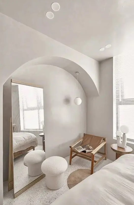 a contemporary bedroom in cold neutral tones, with an arched niche with a floor mirror, a wood and leather chair and a lamp