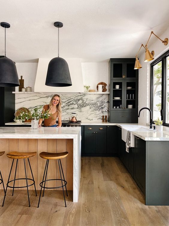 a contrasting kitchen with black cabinets, white marble countertops and a backsplash, a kitchen island, black pendant lamps and gold sconces