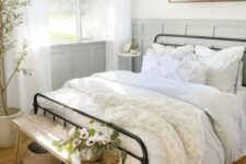 a cute and welcoming modern farmhouse bedroom with grey paneled walls, a wrought bed with neutral bedding, a stained bench and baskets, a potted tree