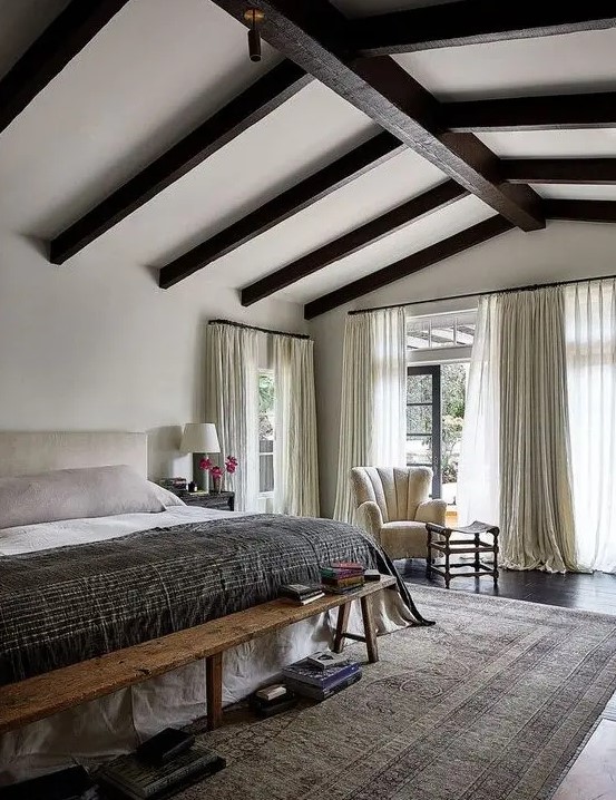 a farmhouse bedroom with dark wooden beams, a neutral bed and a wooden bench, a creamy char, neutral bedidng and a printed rug