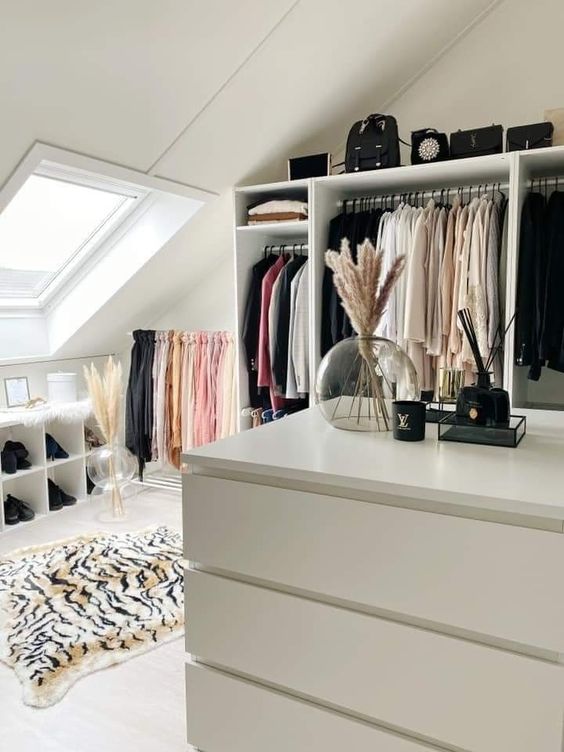 a glam attic closet with an open wardrobe, a storage bench, a dresser, a bold printed rug and some pampas grass