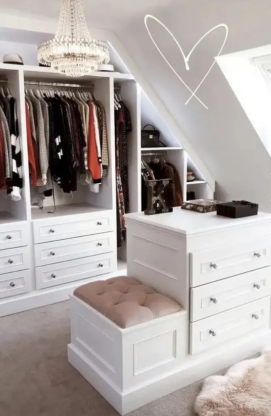a glam sloped closet with open shelves and drawers, a dresser with a stool, a chic crystal chandelier is amazing
