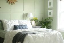 a light green bedroom with a paneled wall, a bed with neutral bedding, a nightstand, potted greenery, a burst mirror