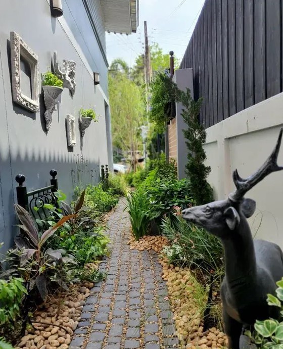 a lovely and creative side yard with a stone path and pebbles, greenery and shrubs, a black forged bench, a black deer and frames and plants on the wall