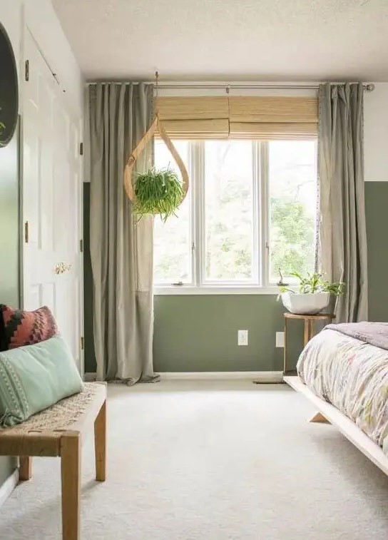 a lovely bedroom with green walls, a bed with printed bedding, a neutral floor and grey curtains, a woven bench and some plants