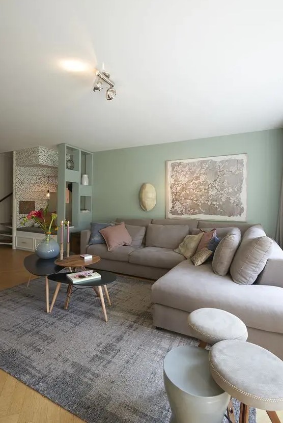a lovely living room with pale green walls, a grey sectional with pillows, an arrangement of coffee tables and stools