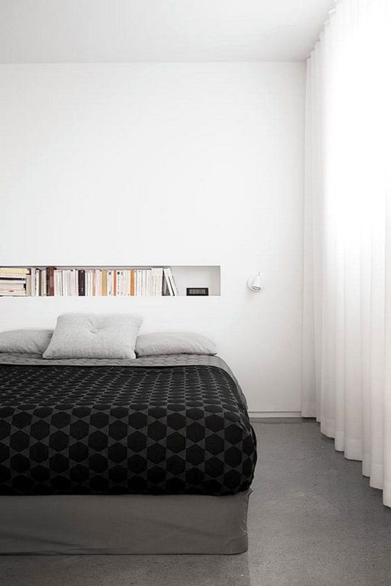 a minimal bedroom with a niche at the headboard that is used as a bookshelf, a bed with monochromatic bedding and a sconce