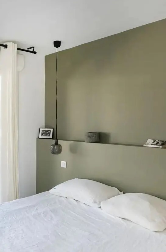 a minimal bedroom with an olive green accent wall, a white bed and bedding, a pendant lamp and some decor