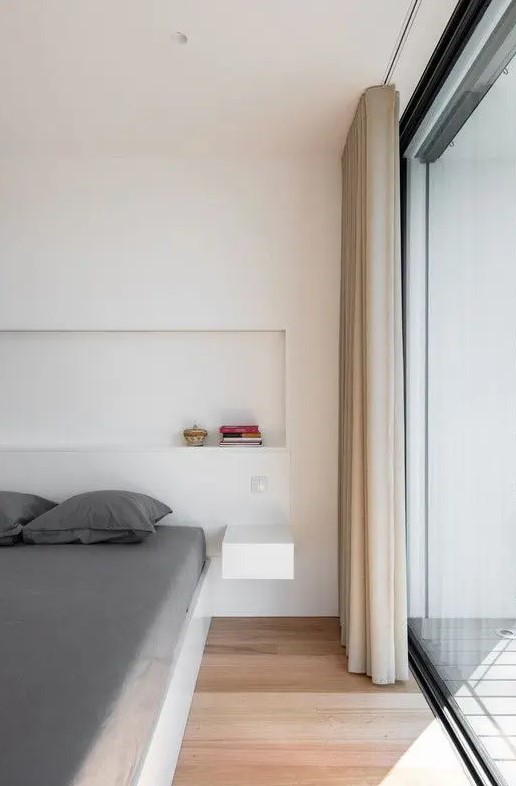 a minimalist bedroom with a glazed wall, a low bed with grey bedding, a niche for decor and a floating nightstand