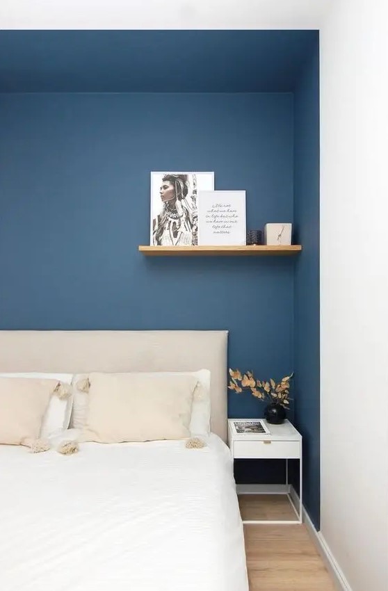 a modern bedroom with a large niche painted navy, a neutral bed with bedding in it, a shelf and a white nightstand