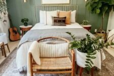 a modern bedroom with a sage green accent wall, a metal bed with neutral bedding, a woven chair, potted plants, gold touches for more elegance