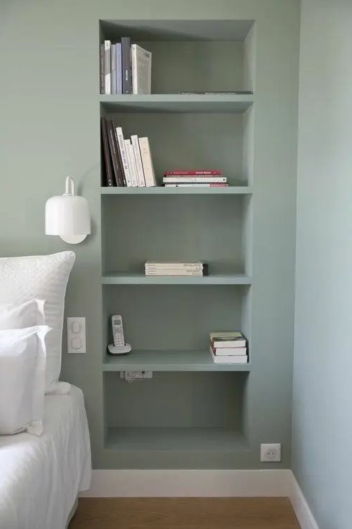 a modern bedroom with olive green walls and niches with books, a white bed with bedding and a white sconce