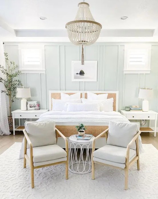 a modern coastal farmhouse bedroom with pale blue paneled walls, a cane bed, pale blue chairs, white nightstands and a bead chandelier