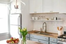 a modern country kitchen with white and light grey cabinets, butcherblock countertops, a shiny subway tile backsplash and a pendant lamp