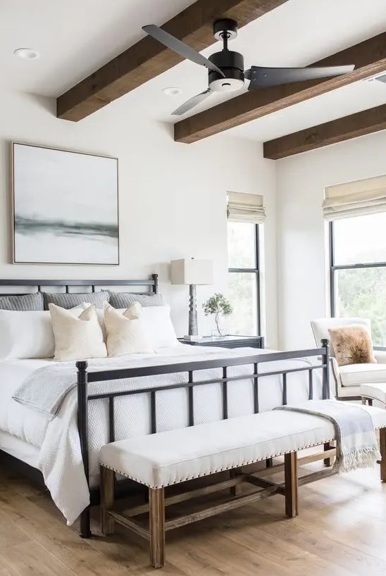 a modern farmhouse bedroom with a black forged bed, a neutral upholstered bench, wooden beams and neutral chairs