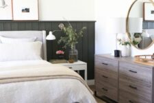 a modern farmhouse bedroom with a dark grene paneled wall, a gray bed with neutral bedding, a stained dresser and a striped rug