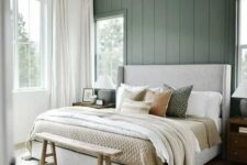a modern farmhouse bedroom with a green accent wall, a grey upholstered bed with neutral bedding, a wooden bench and a chandelier