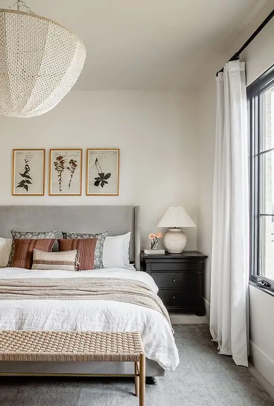 a modern farmhouse bedroom with a grey upholstered bed and neutral bedding, a woven bench, black nightstands and artwork