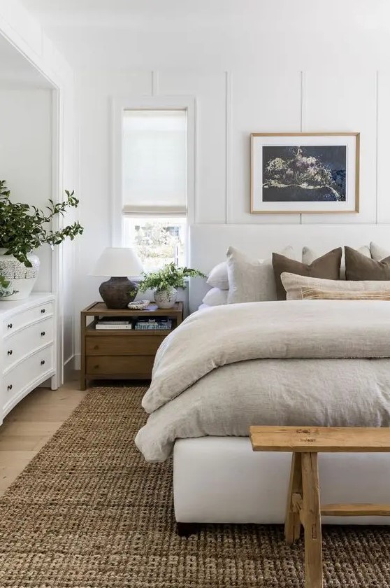 a modern farmhouse bedroom with a white upholstered bed with neutral bedding, a wooden bench, stained nightstands, a white dresser and decor