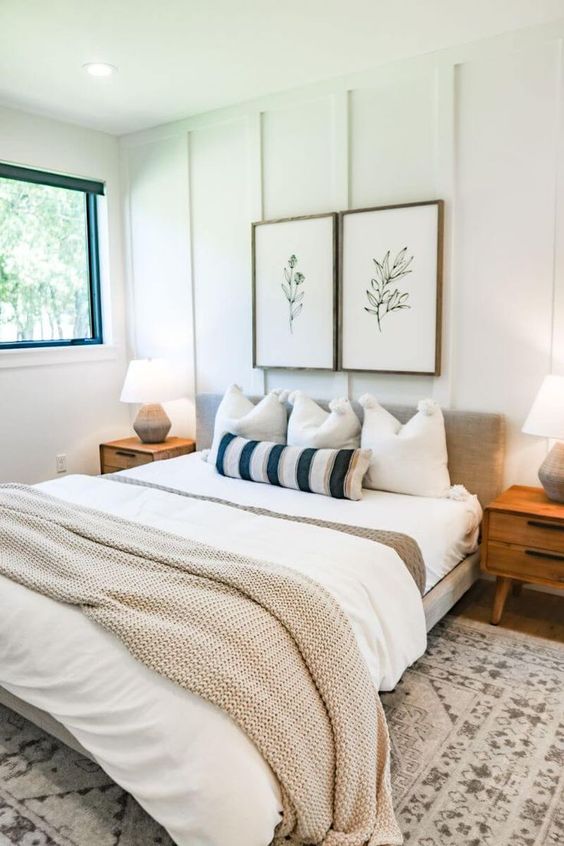 a modern farmhouse bedroom with paneled walls, a neutral bed with layered bedding, stained nightstands and some art