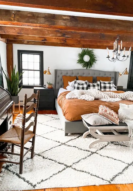 a modern farmhouse meets boho bedroom with stained wooden beams, a grey upholstered bed with bright bedding, black nightstands, a chandelier and greenery