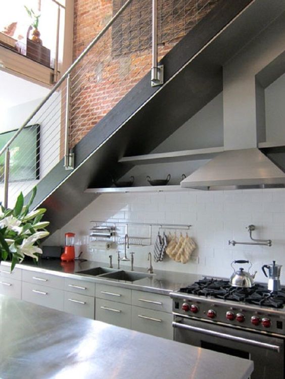 a modern industrial kitchen with metal cabinets and shelves, a hood, a cooker built right under the stairs