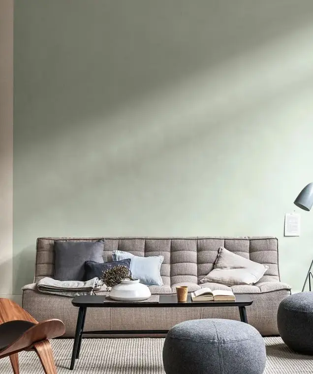 a modern living room with a light green accent wall, a low grey sofa, some poufs, a chair, a floor lamp and a table