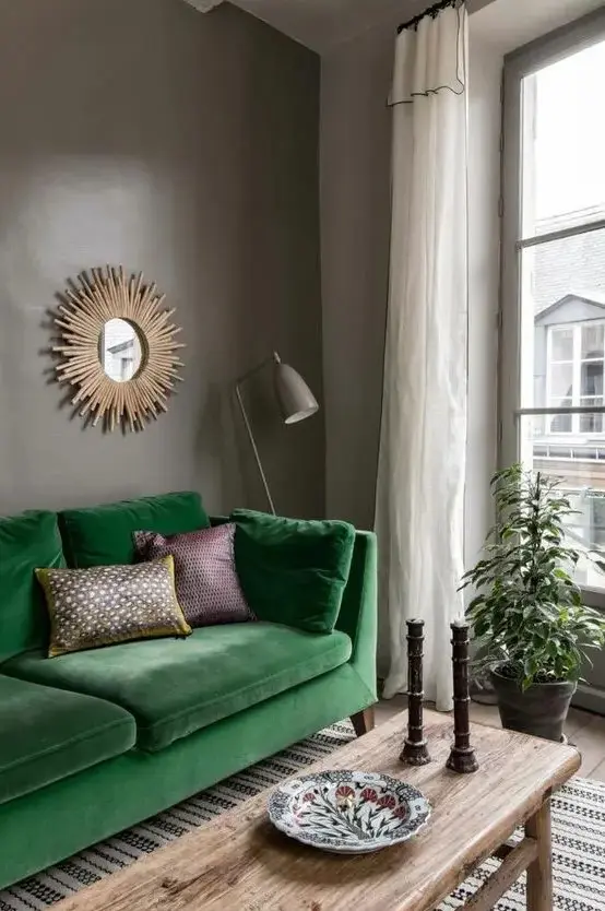 a moody living room with grey walls, a bold green sofa, a stained bench as a coffee table, a burst mirror and a taupe floor lamp