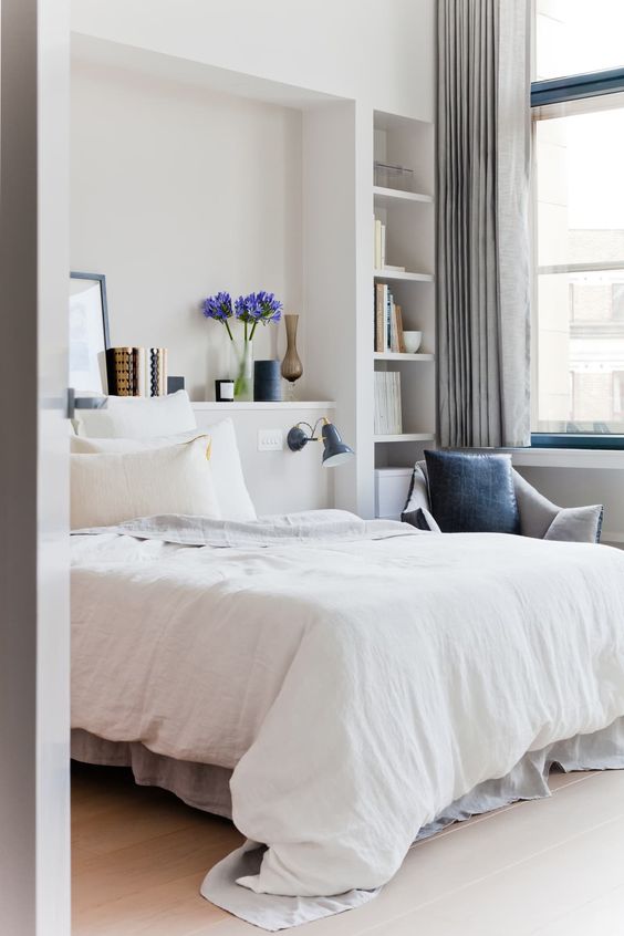 a neutral bedroom with niches, a bed in one of them, others used as shelves, neutral bedding, a navy chair and grey curtains