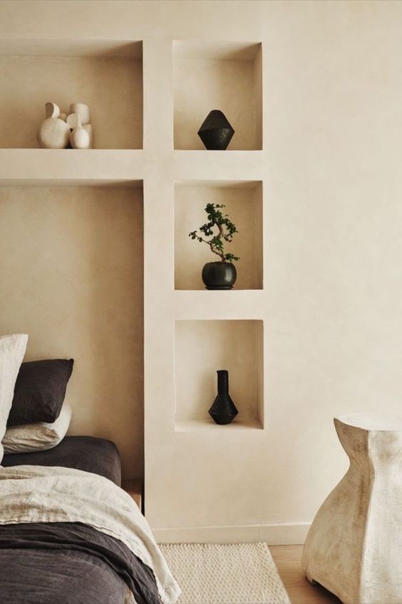 a neutral bedroom with niches, with a bed in one of them, some art and a potted plant plus a sculptural side table