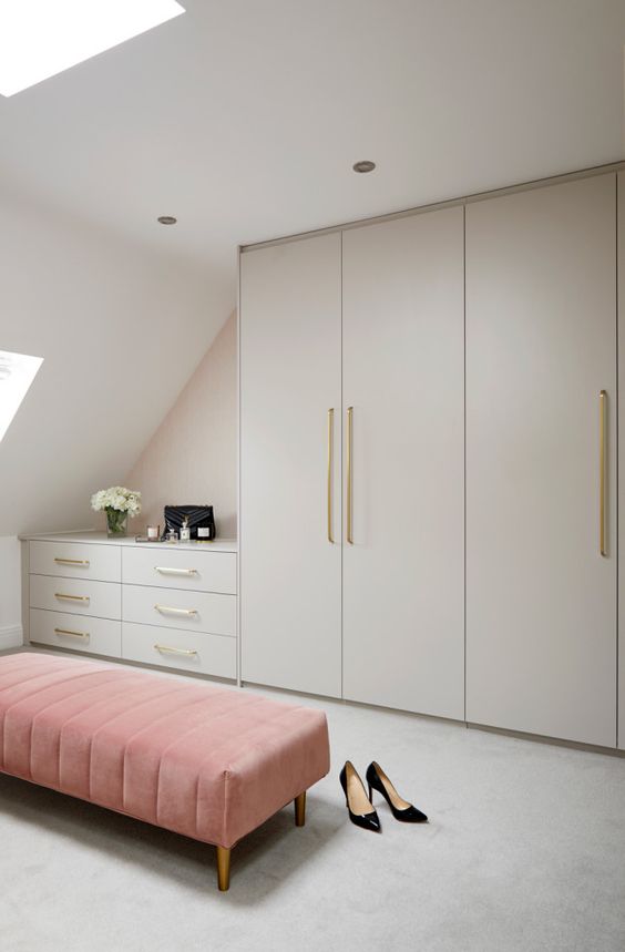 a neutral contemporary attic closet with large wardrobes, dressers, a pink ottoman and skylights to maximize the light
